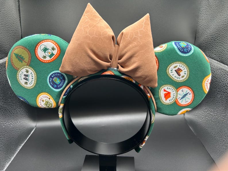 May include: Green fabric mouse ears with a brown bow. The ears are covered in a pattern of colourful patches with text like "Let's Go On A Hike" and "Happy Camper".