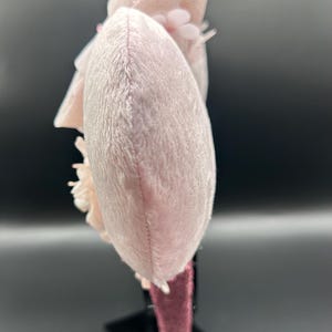 Op de afbeelding: Een lichtroze pluche haarband met een afgeronde oorschelpvorm en een zachte, pluizige textuur. Het accessoire is versierd met gelaagde stoffen bloemen in bijpassende lichtroze tinten, bevestigd aan een donkerroze met fluweel bekleed frame.