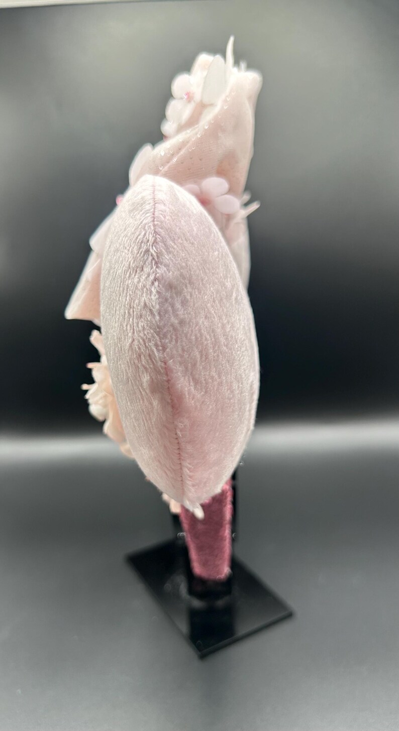 Op de afbeelding: Een lichtroze pluche haarband met een afgeronde oorschelpvorm en een zachte, pluizige textuur. Het accessoire is versierd met gelaagde stoffen bloemen in bijpassende lichtroze tinten, bevestigd aan een donkerroze met fluweel bekleed frame.