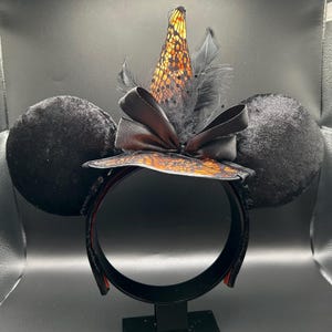 Puede incluir: Una diadema con dos grandes orejas de terciopelo negro y un sombrero de bruja central. El sombrero es de tela naranja con encaje negro, decorado con un lazo de sat&eacute;n negro y plumas negras. La estructura de la diadema es negra con lentejuelas.