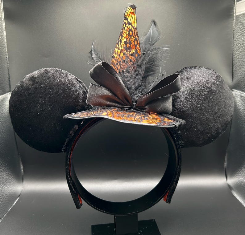 Puede incluir: Una diadema con dos grandes orejas de terciopelo negro y un sombrero de bruja central. El sombrero es de tela naranja con encaje negro, decorado con un lazo de sat&eacute;n negro y plumas negras. La estructura de la diadema es negra con lentejuelas.