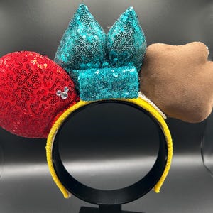 May include: A yellow headband with a red sequined ear, a teal sequined bow, and a brown paw-shaped accent. The red ear has a small cluster of rhinestones. The headband is a fun accessory.