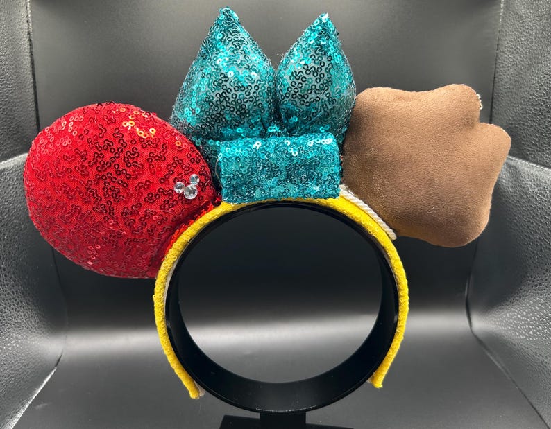 May include: A yellow headband with a red sequined ear, a teal sequined bow, and a brown paw-shaped accent. The red ear has a small cluster of rhinestones. The headband is a fun accessory.
