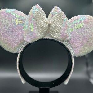 Puede incluir: Orejas de Minnie Mouse blancas con lentejuelas iridiscentes y un lazo adornado con perlas.