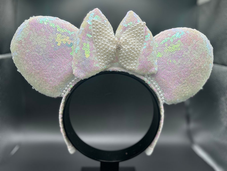 Puede incluir: Orejas de Minnie Mouse blancas con lentejuelas iridiscentes y un lazo adornado con perlas.