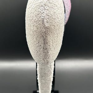May include: A decorative headband featuring a white sequined design. The headband has a curved shape and is adorned with a pink and blue sequined accent. A brown, fuzzy accent is also visible.
