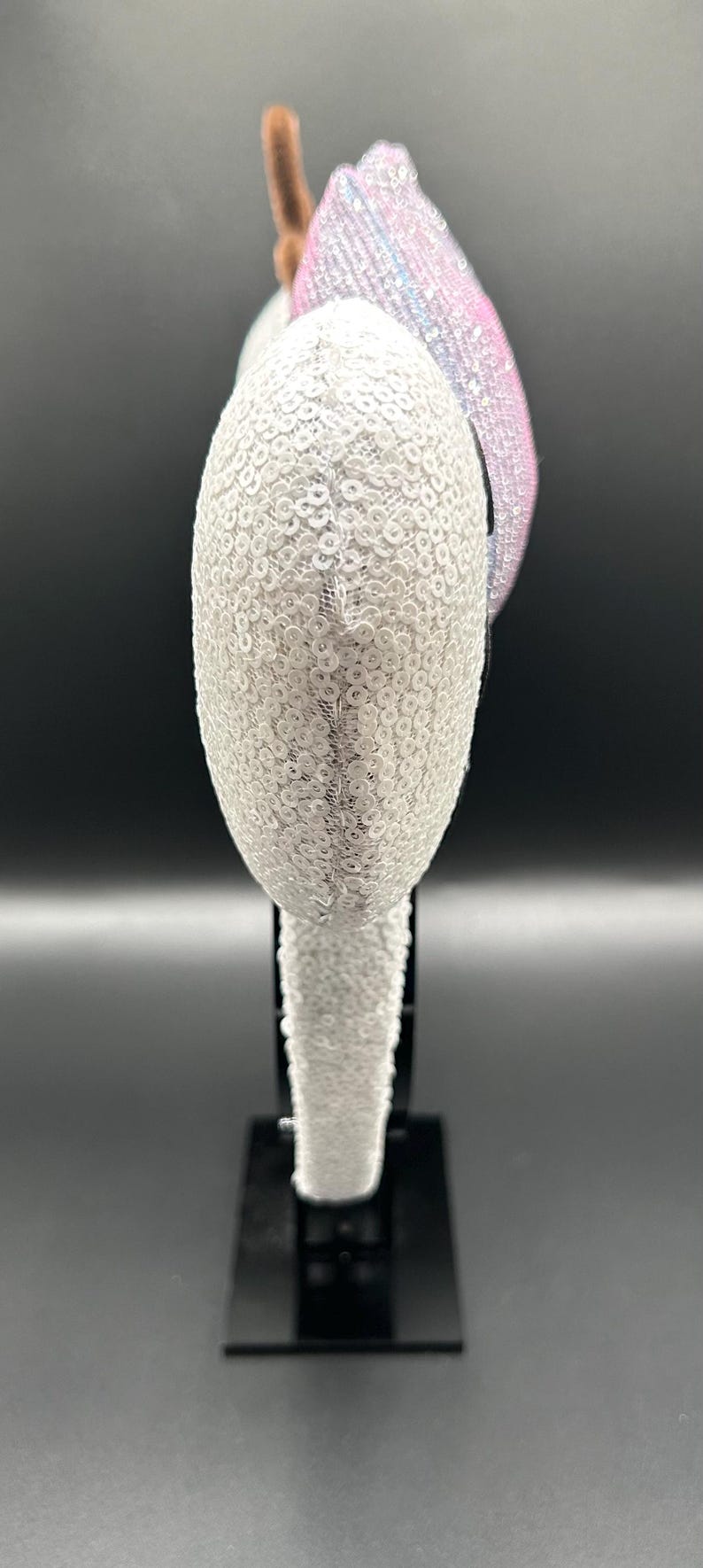 May include: A decorative headband featuring a white sequined design. The headband has a curved shape and is adorned with a pink and blue sequined accent. A brown, fuzzy accent is also visible.