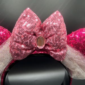 Op de afbeelding: Roze pailletten Minnie Mouse oren met een witte tule strik en een heldere strass in het midden.