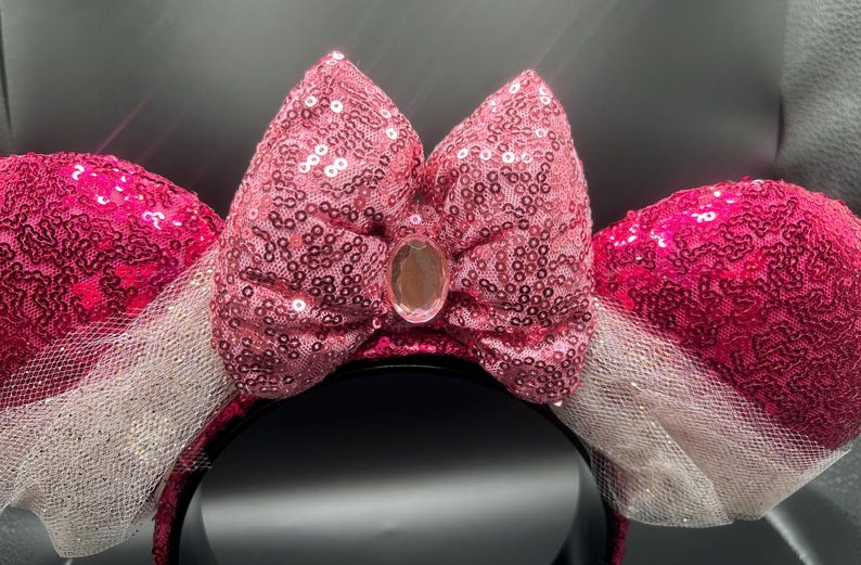 Op de afbeelding: Roze pailletten Minnie Mouse oren met een witte tule strik en een heldere strass in het midden.