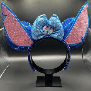 May include: Blue sequined headband with large, glittery pink and blue ears. A blue sequined bow features a Stitch character embellishment. The headband is displayed on a black stand.