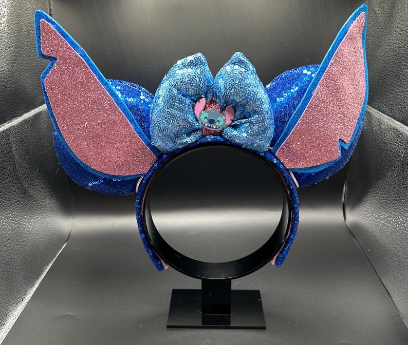 May include: Blue sequined headband with large, glittery pink and blue ears. A blue sequined bow features a Stitch character embellishment. The headband is displayed on a black stand.
