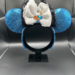 May include: Blue sequined mouse ears with a white bow, black sequined accents, and a character pin. The headband is black, and the ears are round. The bow has a white lace trim.