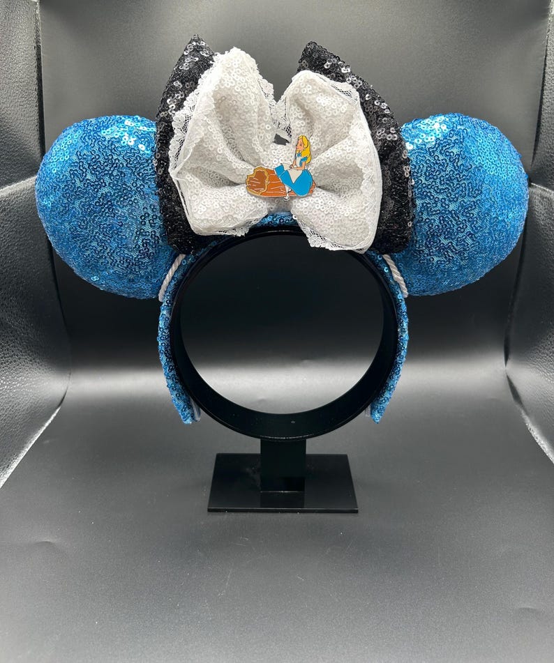 May include: Blue sequined mouse ears with a white bow, black sequined accents, and a character pin. The headband is black, and the ears are round. The bow has a white lace trim.
