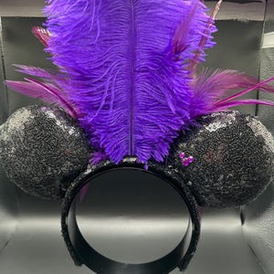 May include: A headband featuring two round ears covered in black sequins, accented with a cluster of three small pink rhinestones on the right ear. A large, vibrant purple ostrich feather plume stands upright between the ears, flanked by smaller dark purple feathers.
