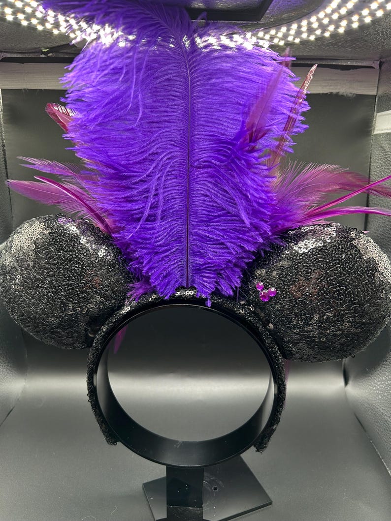 May include: A headband featuring two round ears covered in black sequins, accented with a cluster of three small pink rhinestones on the right ear. A large, vibrant purple ostrich feather plume stands upright between the ears, flanked by smaller dark purple feathers.