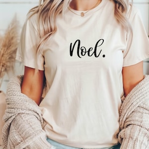 Puede incluir: Una camiseta blanca con la palabra "Noël" escrita en cursiva negra.