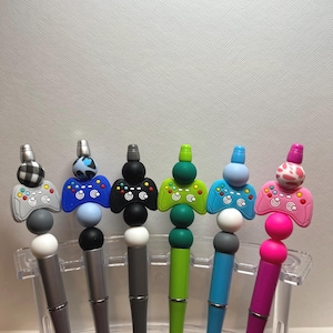 May include: Seven pens with different colored beads and a game controller charm on top. The pens are silver with black ink.