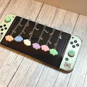 Peut inclure: Une console Nintendo Switch avec un design vert clair et blanc, avec un motif d'ours et de feuilles. Six breloques avec des nuages pastel, des chaînes argentées, des lunes et des étoiles sont attachées à l'écran. La console repose sur une surface en bois.