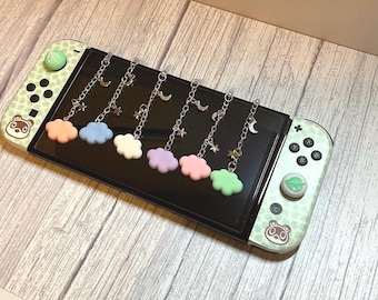 May include: A Nintendo Switch console with a light green and white design, featuring a bear and leaf pattern. Attached to the screen are six charms with pastel cloud shapes, silver chains, moons, and stars. The console rests on a wooden surface.