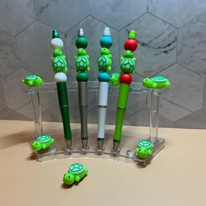 May include: Five decorative pens with turtle designs. The pens have green, silver, and white barrels, with turtle and bead accents. A clear acrylic pen holder displays the pens, with additional turtle figurines.