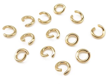 May include: A group of 12 gold jump rings, scattered on a white background. The jump rings are all the same size and shape, and they are made of a shiny metal.