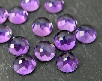 Pode incluir: Uma coleção de 12 gemas de ametista facetadas, cada uma com uma forma redonda e um tom roxo. As gemas estão dispostas em um grupo sobre uma superfície cinza escura.