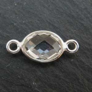 Op de afbeelding: Zilveren connector met een helder ovaalvormig kristal. De connector heeft aan beide zijden twee lussen.