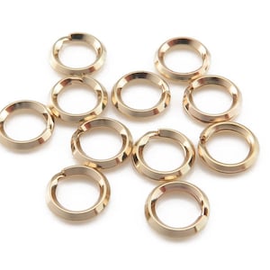 May include: Ten gold jump rings, each with a slightly rounded, textured edge. The rings are arranged in a loose cluster on a white background.