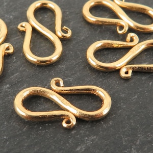 Może przedstawiać: Sześć złotych metalowych zapięć w kształcie litery 'S' o gładkim, polerowanym wykończeniu. Zapięcia są ułożone w półkole na ciemnoszarym tle.