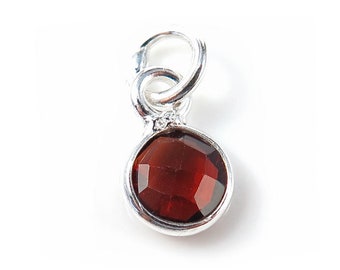 Puede incluir: Un charm de plata con una piedra preciosa roja oscura, redonda y facetada. El charm tiene un lazo en la parte superior para sujetarlo a una pulsera o collar. La piedra está engastada en un bisel de plata.