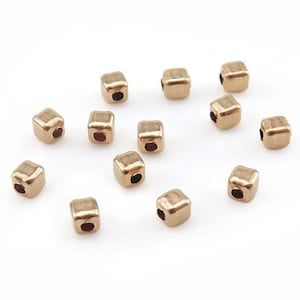 Puede incluir: Cuentas doradas en forma de cubo, con bordes redondeados y un pequeño agujero en el centro. Las cuentas están dispersas sobre una superficie blanca, ideales para la creación de joyas o manualidades.