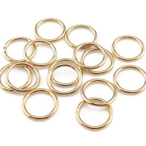 Puede incluir: Un grupo de 12 anillos de salto dorados, todos del mismo tamaño, dispuestos en una pila suelta sobre un fondo blanco.