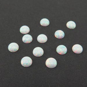 Puede incluir: Una colección de ópalos redondos tallados en cabujón. Los ópalos son blancos con destellos iridiscentes de color, incluyendo rosa, azul y verde. Están dispersos sobre una superficie gris oscuro. Estas piedras preciosas son adecuadas para la fabricación de joyas.