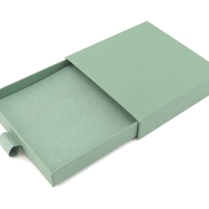 May include: A pale green rectangular gift box with a sliding lid, partially open to reveal the interior. The box has a textured finish and a small tab for opening. The box is set against a white backdrop.