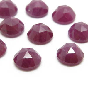 May include: Ten faceted ruby gemstones, each with a deep red colour and a polished finish. The gemstones are arranged in a scattered pattern on a white surface.