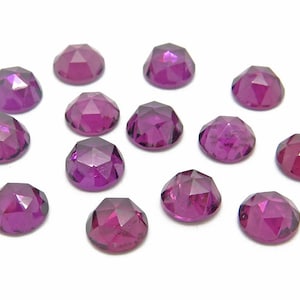 May include: A collection of 13 purple gemstones, each with a faceted cut, arranged on a white surface.