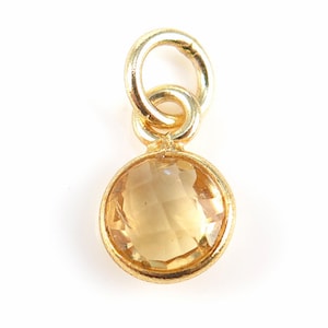 May include: A gold pendant featuring a round, faceted, light-yellow gemstone. The pendant has a small loop at the top for attaching to a chain or bracelet. The gemstone is set in a gold bezel.