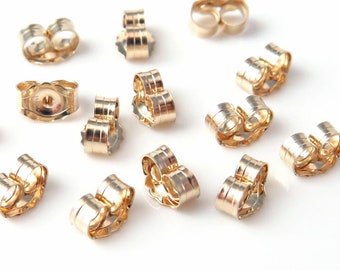 May include: A close-up of a group of gold-toned earring backs. The backs are designed to fit over the post of an earring and have a small, round hole in the centre.