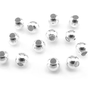 Puede incluir: Un conjunto de cuentas metálicas brillantes de color plateado. Cada cuenta es esférica con un pequeño agujero gris oscuro visible en un lado. Las cuentas están dispersas sobre un fondo blanco, para la creación de joyas o artesanías.