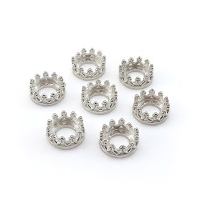 Puede incluir: Siete hallazgos de joyería en forma de corona de color plateado. Cada corona tiene una base circular con un diseño detallado y ornamentado. Las coronas están dispuestas sobre un fondo blanco, mostrando sus intrincados detalles y brillo metálico.