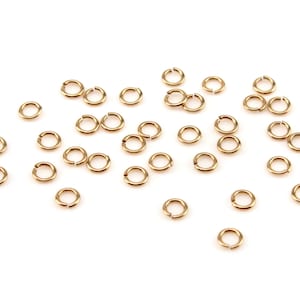 May include: A scattering of small, gold-coloured jump rings on a white background. These open rings are used in jewellery making and crafts. The circular rings have a small opening for connecting other components.