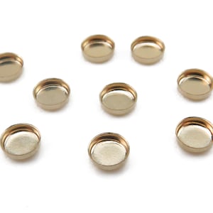 May include: Ten gold-colored metal round bezel settings for jewelry making. The bezels are all the same size and are arranged in a circle on a white background.