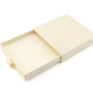 Puede incluir: Una caja rectangular abierta, de color crema, con tapa corredera. La caja está vacía, revelando un interior de color claro. Una pequeña pestaña enrollada es visible en el lateral de la caja. La caja está sobre un fondo blanco.