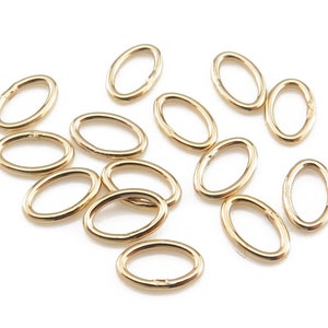May include: A selection of small, gold-coloured oval jump rings scattered on a white surface. These jewellery findings are open and ready for use in various crafting projects. The rings are made of a shiny, metallic material.
