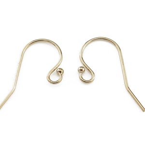 Puede incluir: Dos ganchos dorados para pendientes sobre un fondo blanco. Cada gancho tiene una parte superior curvada con una pequeña bola en el extremo y una parte inferior recta y puntiaguda. Los ganchos están diseñados para la fabricación de joyas.