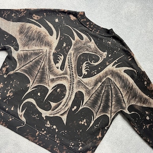 May include: Black sweatshirt with a white dragon graphic print. The dragon has wings and a long tail. The sweatshirt has a distressed look with white splatter paint.