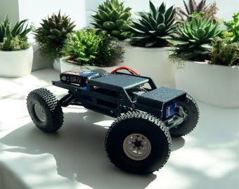 May include: A black and gray remote control rock crawler with large, knobby tires. The vehicle has a detailed chassis and is positioned in front of a row of potted succulents. The tires are approximately 4 inches in diameter.