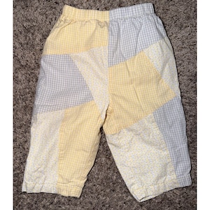 May include: Pants featuring a patchwork design with alternating panels of yellow gingham, light gray gingham, and a white fabric with a small yellow floral print. The garment has an elastic waistband and a straight-leg cut with visible seams.