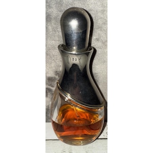 May include: A vintage perfume bottle with a silver-colored cap and neck. The clear glass bottle contains a golden-brown liquid. The bottle has a unique shape, with a rounded base and a slender neck. A thin, gold-colored band wraps around the bottle.