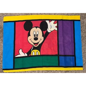 Könnte beinhalten: Ein rechteckiger Kissenbezug mit Micky Maus in einem farbenfrohen, geometrischen Design. Micky wird mit einem lächelnden Gesichtsausdruck dargestellt, der mit der Hand winkt. Der Kissenbezug hat einen gelben Rand mit blauen, roten, grünen und violetten Abschnitten.